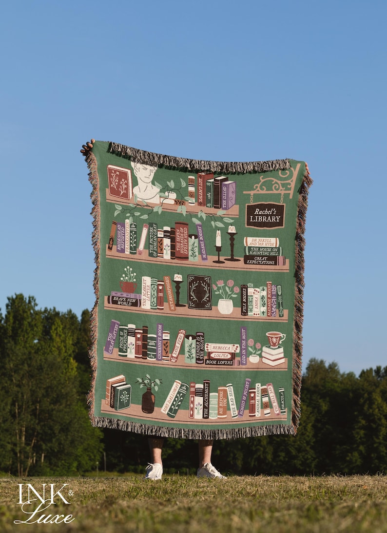 May include: A green woven throw blanket with a bookshelf design featuring a variety of book titles. The blanket has a fringe border and the text "Rachel's Library" is displayed on a sign hanging from the top shelf.