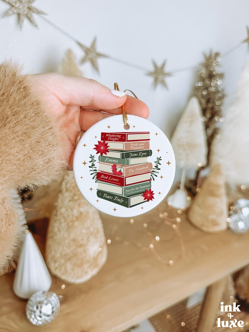 May include: A round white ceramic ornament featuring a stack of colourful books with titles including Wuthering Heights, Pride and Prejudice, Jane Eyre, Emma, The Notebook, Book Lovers, Just for the Summer, and Yours Truly, accented by red flowers.