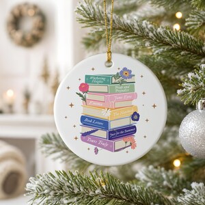 May include: A round white ceramic ornament featuring a stack of colorful books with titles including Wuthering Heights, Pride and Prejudice, Jane Eyre, Emma, The Notebook, Book Lovers, Just for the Summer, and Yours Truly, accented by small floral illustrations.