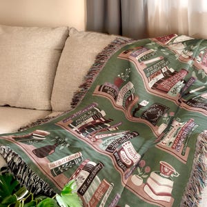 May include: A green and pink woven throw blanket with a bookshelf design featuring the titles of classic novels. The blanket has a fringe border.