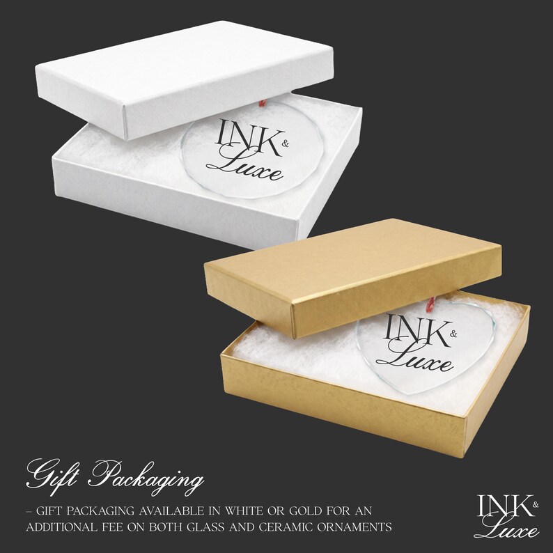 May include: Two rectangular gift boxes, one white and one gold, each containing a clear, circular or heart-shaped glass ornament. The ornaments feature the black text INK & LUXE. The boxes are lined with white padding for the ornaments.