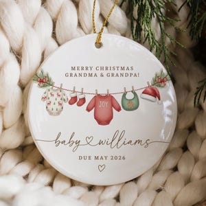 May include: A round white ceramic ornament featuring a festive clothesline illustration with a baby onesie, red socks, a red shirt with the word JOY, a green bib with a gingerbread man, and a Santa hat. Text reads MERRY CHRISTMAS GRANDMA & GRANDPA!, baby williams, and DUE MAY 2026.