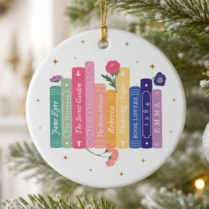 May include: A round white ceramic ornament with a gold hanging string, featuring a colourful illustration of a row of books with titles including Jane Eyre, The Notebook, The Secret Garden, Pride & Prejudice, The Secret History, Rebecca, Wuthering Heights, Book Lovers, 1984, and Emma, accented with small floral and star motifs.