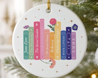 May include: A round white ceramic ornament with a gold hanging string, featuring a colorful illustration of a row of books with titles including Jane Eyre, The Notebook, The Secret Garden, Pride & Prejudice, The Secret History, Rebecca, Wuthering Heights, Book Lovers, 1984, and Emma, accented with small floral and star motifs.