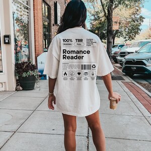 May include: A white t-shirt with a graphic design that says "100% TBR" and "Romance Reader". The graphic design is in a black and white color scheme and includes a barcode and other design elements.