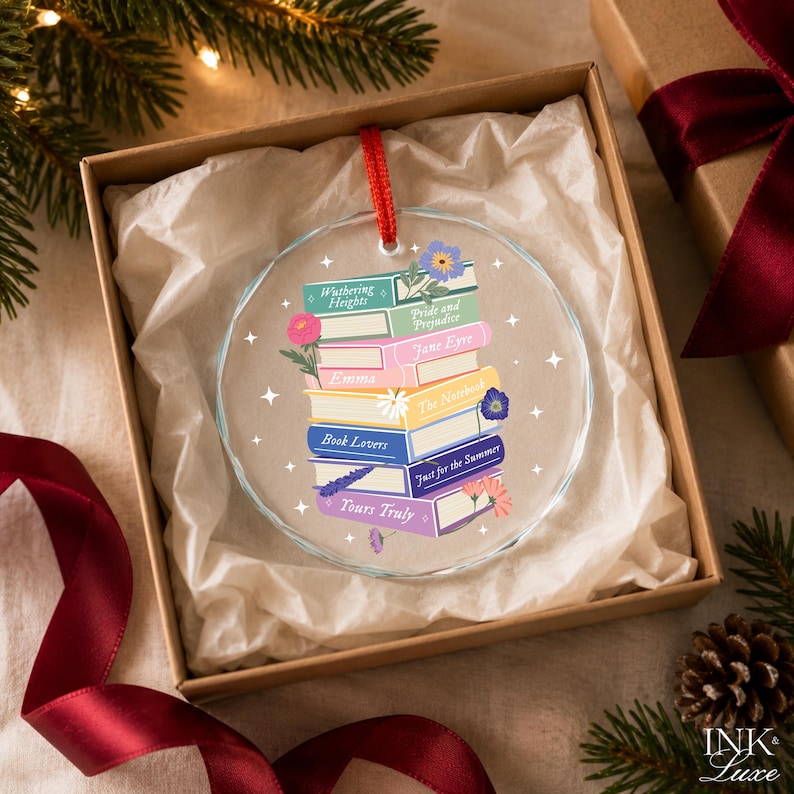 May include: A round, clear glass ornament featuring a stack of colorful books with titles including Wuthering Heights, Pride and Prejudice, Jane Eyre, Emma, The Notebook, Book Lovers, Just for the Summer, and Yours Truly, accented with small floral illustrations.