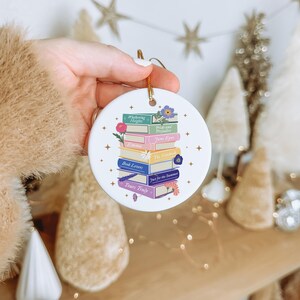 May include: A round white ceramic ornament featuring a stack of colorful books with floral accents. The book spines are labeled with titles including Wuthering Heights, Pride and Prejudice, Jane Eyre, Emma, The Notebook, Book Lovers, and Yours Truly.