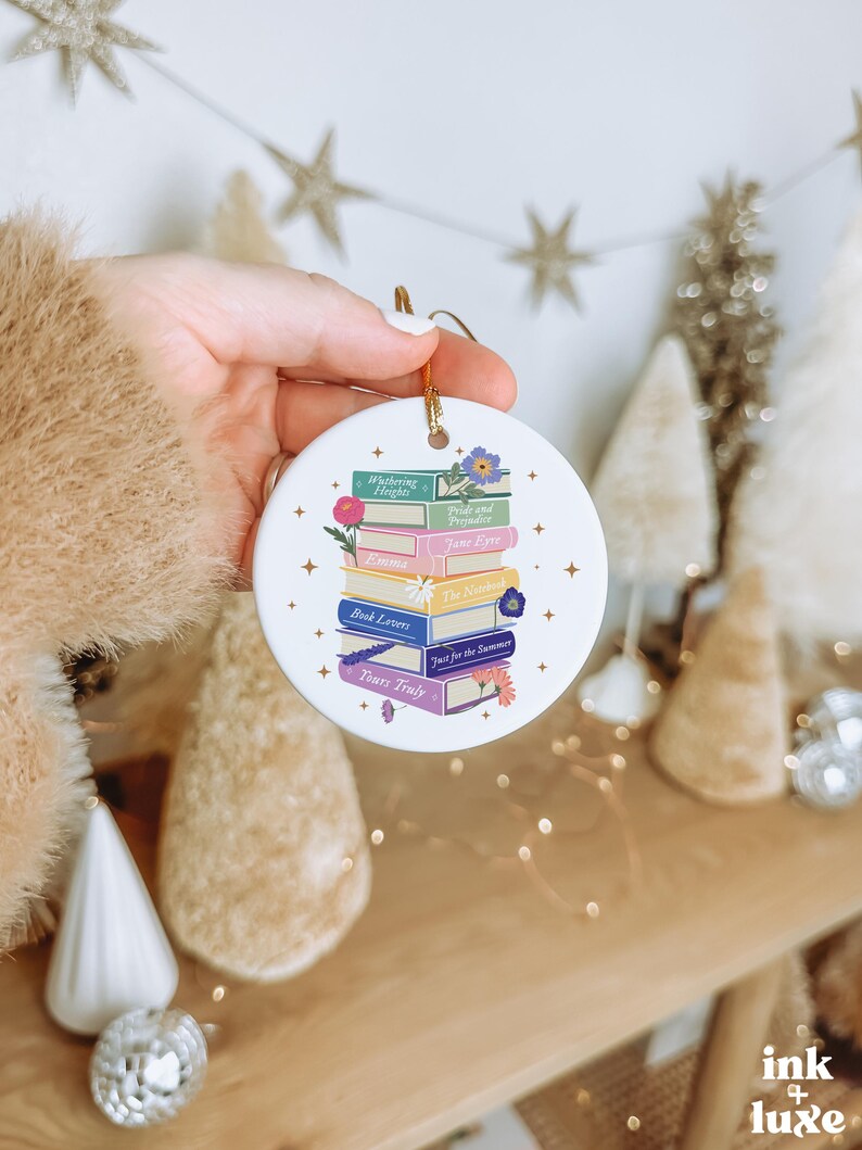 May include: A round white ceramic ornament featuring a stack of colorful books with floral accents. The book spines are labeled with titles including Wuthering Heights, Pride and Prejudice, Jane Eyre, Emma, The Notebook, Book Lovers, and Yours Truly.