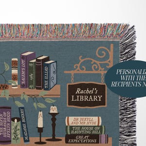 May include: A blue throw blanket with a library theme. It features a bookshelf design with book titles like "The Iliad" and "Madame Bovary." The blanket has a fringe border and a sign that reads "Rachel's Library."