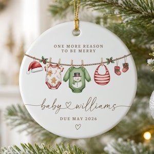 May include: A round white ceramic ornament featuring a clothesline illustration with a Santa hat, a gingerbread-patterned baby onesie, a snowman onesie, a red striped bib, and baby socks. Text reads: ONE MORE REASON TO BE MERRY, baby WILLIAMS, DUE MAY 2026.