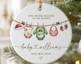 May include: A round white ceramic ornament featuring a clothesline illustration with a Santa hat, a gingerbread-patterned baby onesie, a snowman onesie, a red striped bib, and baby socks. Text reads: ONE MORE REASON TO BE MERRY, baby WILLIAMS, DUE MAY 2026.