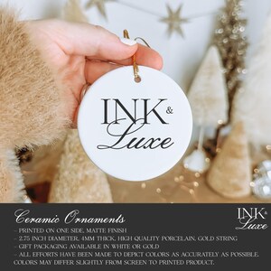 May include: A round white ceramic ornament featuring the black text INK & Luxe printed in the centre. The ornament has a hole at the top with a gold string attached for hanging. The product is 6.985 cm in diameter and 4 mm thick.