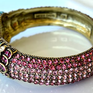 May include: A gold-coloured hinged bangle bracelet adorned with pink, purple, blue, and yellow rhinestones. The bracelet has a large pink gemstone and is decorated with smaller, colourful stones.