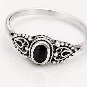 May include: A silver ring with an oval black stone at the center. The ring has an ornate design with filigree and small beaded accents. The ring is set against a white background.