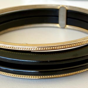 May include: A stack of three black bangles with gold-colored trim. The bangles are stacked together, with a gold-colored band on top. The bangles are on a white surface.