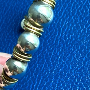 Op de afbeelding: Een zilverkleurige armband met grote, bolvormige kralen. Elke kraal is geaccentueerd met goudkleurig opgerold draad. De armband wordt gepresenteerd tegen een getextureerde, diepblauwe achtergrond.