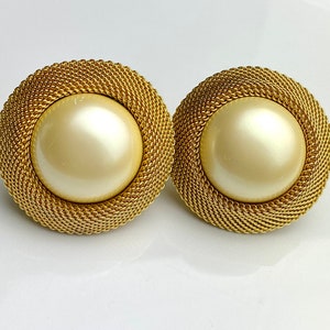 May include: A pair of round, gold-toned clip-on earrings. Each earring features a large, cream-colored, pearl-like center surrounded by a woven gold mesh. The earrings are set against a white background.