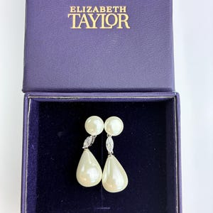May include: A pair of drop earrings featuring a round pearl stud, a small marquise-cut clear stone, and a large teardrop-shaped pearl, presented in a purple Elizabeth Taylor branded jewelry box with a dark blue velvet interior lining.