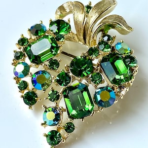 Könnte beinhalten: Vintage Brosche mit herzförmigem Design, verziert mit smaragdgrünen und irisierenden Strasssteinen. Die Brosche hat eine goldfarbene Metalleinfassung mit strukturiertem Blattakzent. Die Edelsteine sind in verschiedenen Formen und Größen.