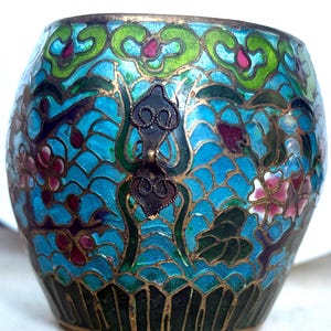 May include: A small, round, decorative cloisonné vase with a turquoise background and floral patterns in pink, green, and gold. The vase has a dark green base and a decorative metal handle.
