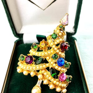 May include: A gold-tone Christmas tree brooch featuring a swirl design accented with small white faux pearls and multi-colored faceted rhinestones in shades of red, blue, green, purple, and yellow, topped with a clear stone and a single pearl base.