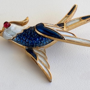 Op de afbeelding: Een goudkleurige vogelbroche met een rood oog en een gedetailleerd ontwerp. Het lichaam van de vogel is diepblauw met witte en zwarte accenten op de vleugels. De broche is waarschijnlijk gemaakt van metaal en email.
