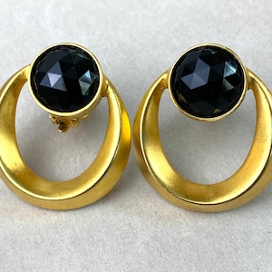 Puede incluir: Un par de pendientes dorados de clip. Cada pendiente presenta una gran gema negra facetada engastada en un marco circular. El marco tiene un acabado liso y mate y una forma de lágrima.