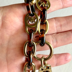 May include: A gold-tone chain bracelet with tortoiseshell links. The bracelet features oval and circular links, with the initials "MK" on some links. The bracelet has a toggle clasp closure.