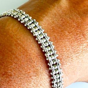Può includere: Bracciale argentato con un design dettagliato a perline. Il bracciale presenta file di piccole perline rotonde collegate da delicati collegamenti. Il gioiello è mostrato sul polso di una persona, evidenziando l'intricata lavorazione e la consistenza dell'accessorio.