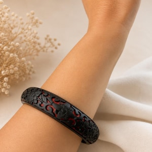 May include: A black bangle bracelet featuring an intricate openwork floral carving that reveals a vibrant red interior layer. The circular accessory has a smooth, matte finish and a continuous pattern of detailed blossoms and leaves around the band.