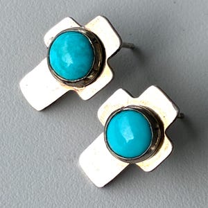 May include: A pair of silver-toned stud earrings. Each earring features a bright turquoise stone set in a silver bezel, mounted on a cross-shaped backing. The earrings are displayed on a gray surface.