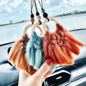 May include: Three wooden ring car air fresheners with tassels. The tassels are orange, blue, and brown. The rings are hanging from a black string with brown and white beads.