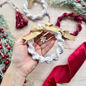 May include: Three handmade macrame Christmas ornaments with a snowflake charm. The ornaments are white, red, and cream with gold ribbon.