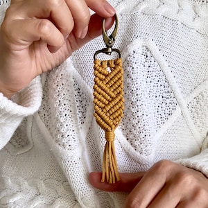 May include: A mustard yellow macrame keychain with a tassel and a silver metal clasp.