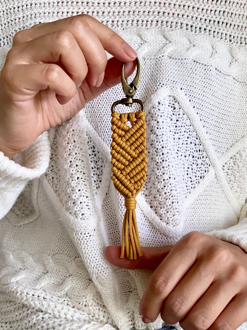 May include: A mustard yellow macrame keychain with a tassel and a silver metal clasp.
