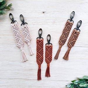 May include: Set of six macrame keychains with tassels. Two are brown, two are beige, and two are a darker brown. The keychains are made with a chevron pattern.