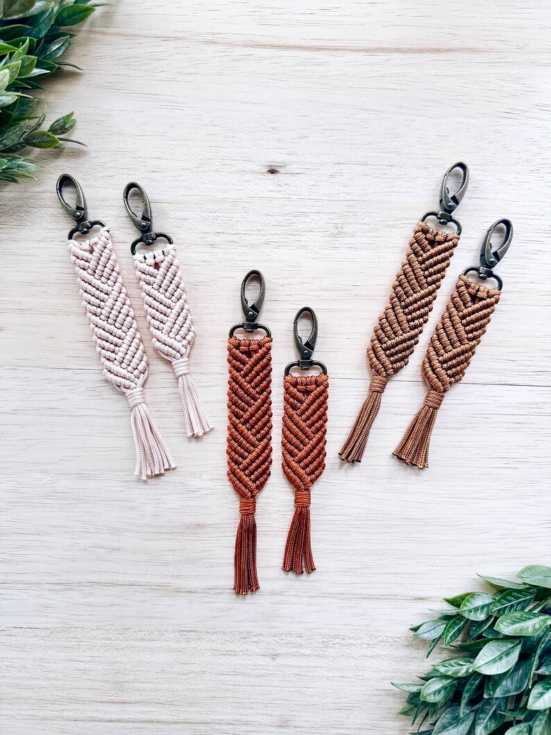 May include: Set of six macrame keychains with tassels. Two are brown, two are beige, and two are a darker brown. The keychains are made with a chevron pattern.