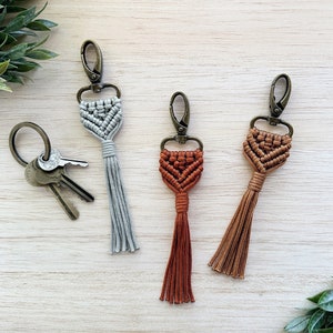 May include: Three keychains with macrame tassels. The keychains are made with a silver metal clasp and have a geometric pattern. One keychain has a grey tassel, one has a rust-coloured tassel, and one has a brown tassel.