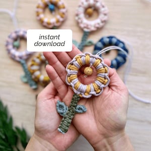 May include: Handmade macrame flower with yellow, gray, and white accents, held in a person's hands. The flower has a green stem and leaves. The text "instant download" is visible in the image. Other similar flowers are in the background.