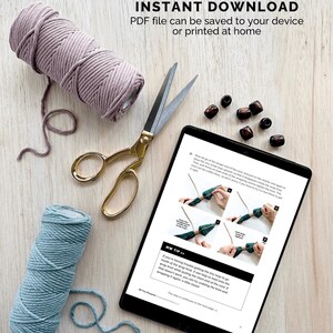 May include: A tablet displaying a digital download pattern for a macrame project. The pattern includes instructions and images. The tablet is on a wooden surface with two spools of cord, a pair of scissors, and wooden beads.