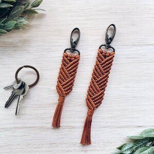 May include: Two brown macrame keychains with tassels and silver metal clasps. The keychains are hanging on a white wooden surface.