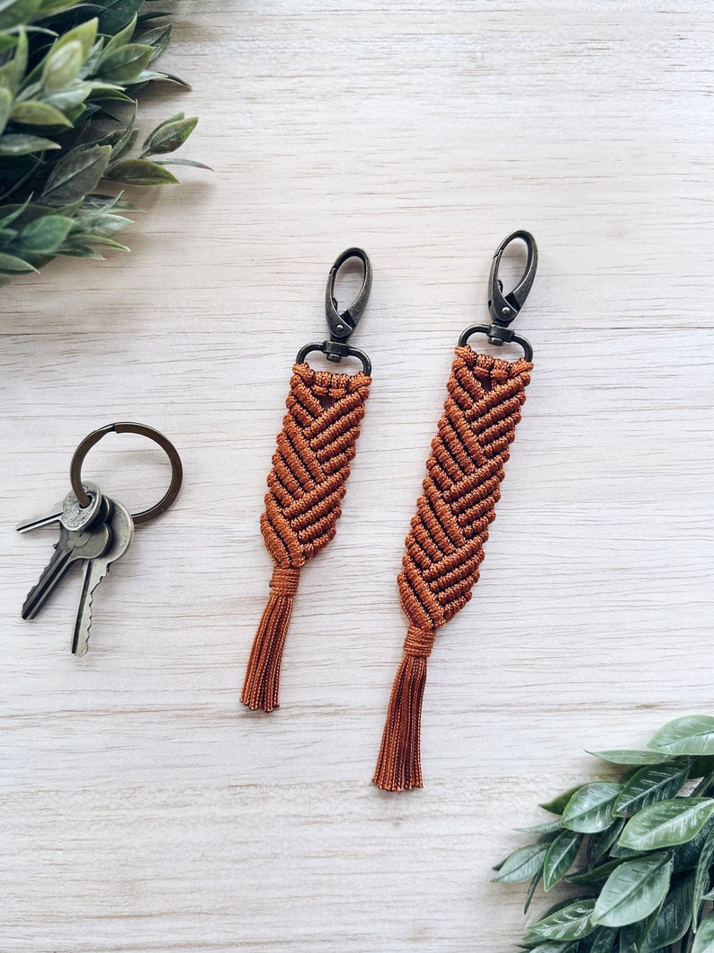 May include: Two brown macrame keychains with tassels and silver metal clasps. The keychains are hanging on a white wooden surface.