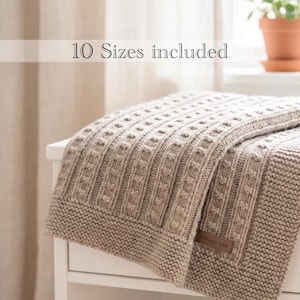 May include: A beige knitted blanket featuring a textured cable pattern and a garter stitch border. A small brown rectangular tag with the text Knit with Love is attached to the corner. The image includes the text 10 Sizes included.