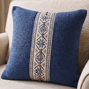 May include: A square blue knit throw pillow featuring a central vertical band of cream-colored fabric. The band displays a blue and gold geometric floral pattern and is embellished with a single row of small, round, light brown wooden beads.