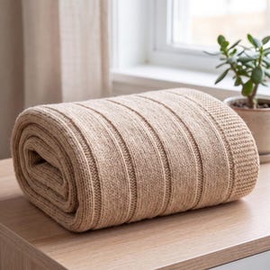 May include: A folded beige knit throw blanket featuring a vertical ribbed texture and a wide, textured border. The fabric has a subtle speckled appearance throughout the weave, creating a consistent neutral tone across the entire piece.