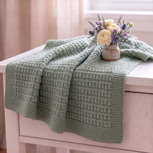 May include: A sage green knitted blanket with a textured waffle stitch pattern and a garter stitch border. A small ceramic vase holds a bouquet of white ranunculus flowers and purple thistle stems, resting on the folded fabric surface.