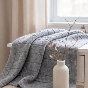 May include: A light gray knitted throw blanket with a textured pattern of horizontal stripes and a garter stitch border. The blanket is draped over a surface next to a white ceramic vase containing a thin, bare, brown branch.