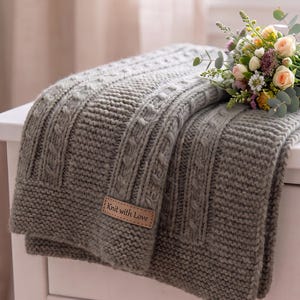 May include: A sage green knitted throw blanket featuring a textured cable knit pattern and a rectangular tan leather label stitched to the corner with the text Knit with Love. A small bouquet of flowers rests on the folded fabric.