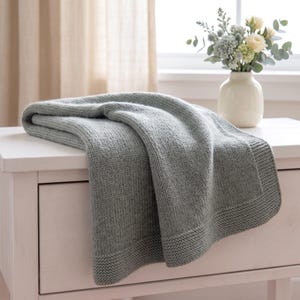 May include: A sage green knitted blanket featuring a textured garter stitch border and a smooth stockinette stitch center. The blanket is folded and draped, showcasing the soft yarn construction and the consistent light green color throughout.