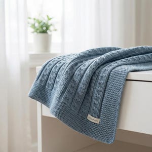 May include: A dusty blue knitted blanket featuring a textured cable knit pattern and a garter stitch border. A small rectangular fabric label is sewn onto the corner of the blanket with the text Knit with Love printed in a dark brown font.